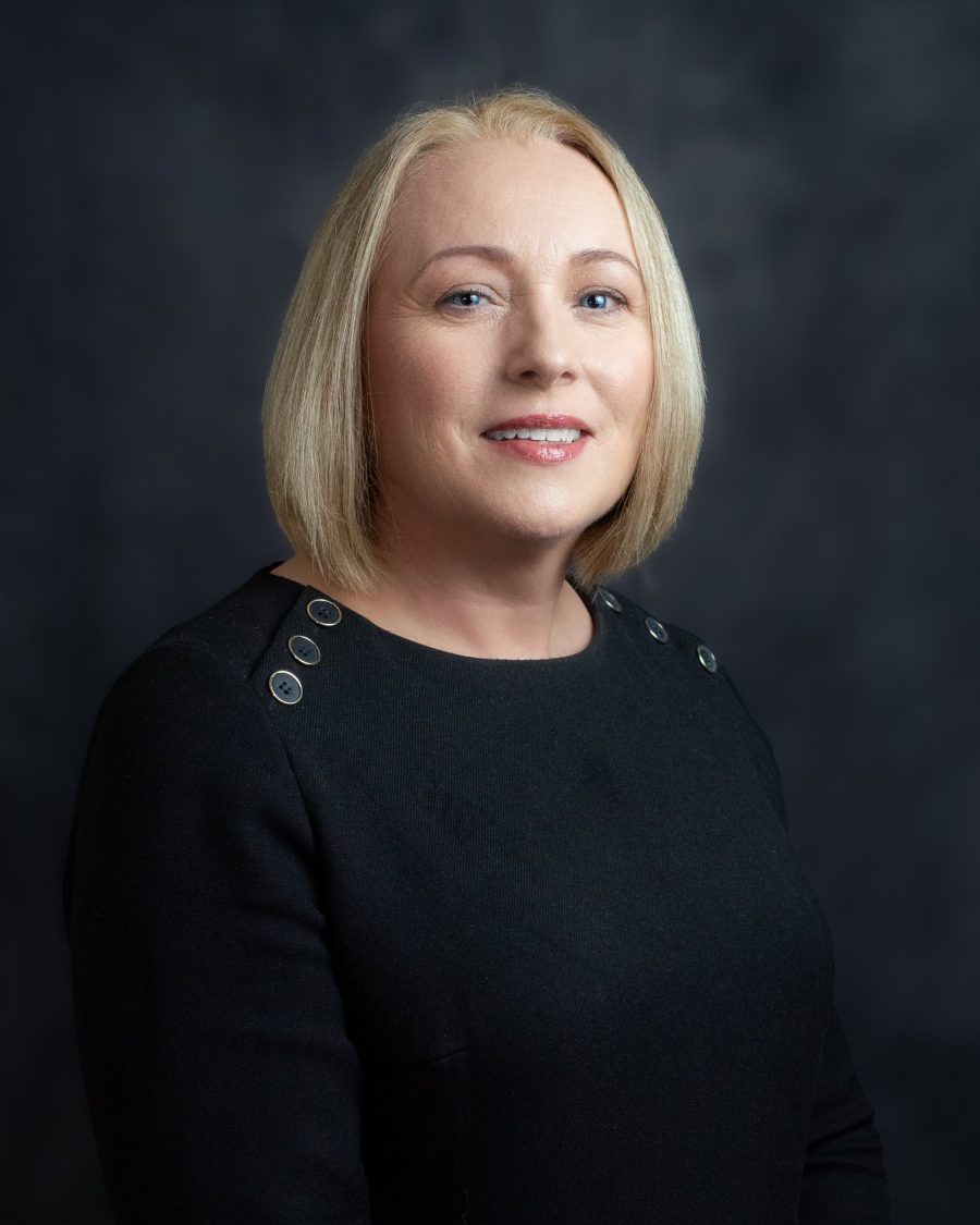 Studio headshot of a woman with a blonde bob in a black top on a dark grey backdrop
