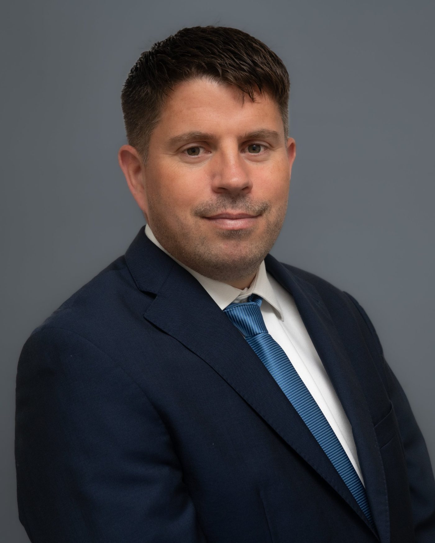 Corporate headshot of a man in a navy suit and blue tie on a blue-grey backdrop
