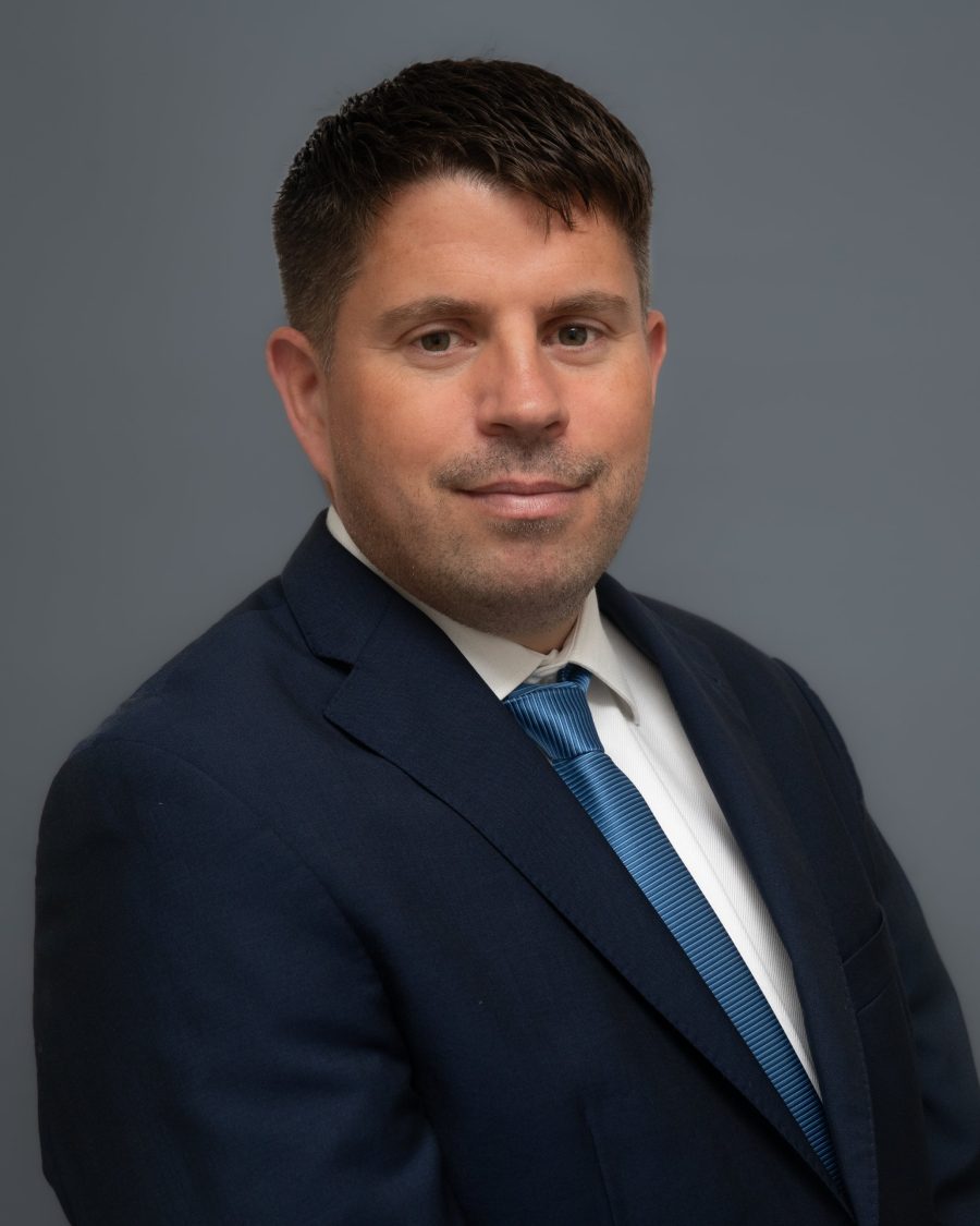 Corporate headshot of a man in a navy suit and blue tie on a blue-grey backdrop