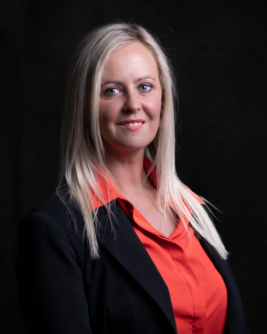 Professional headshot of a woman in a black blazer and red blouse on a black backdrop